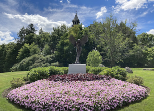 A statue with wings stands in a flower bed of pink blooms, surrounded by lush greenery and a blue sky.