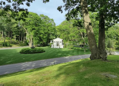 A white house surrounded by lush green trees and a well-manicured lawn, with a road in the foreground.