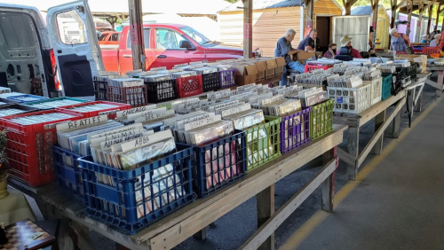 A market stall with wooden tables filled with crates of vinyl records, surrounded by shoppers and vehicles in the background.