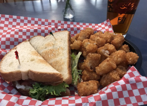 A sandwich with lettuce and bacon, served with a side of crispy tater tots, on a checkered paper plate and a beer.