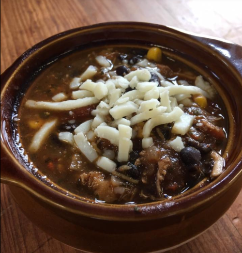 A bowl of hearty soup topped with shredded cheese, featuring beans, corn, and chunks of meat.
