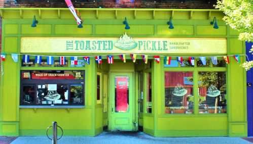 Bright green storefront of "The Toasted Pickle," featuring a sign for handcrafted sandwiches and colorful bunting.