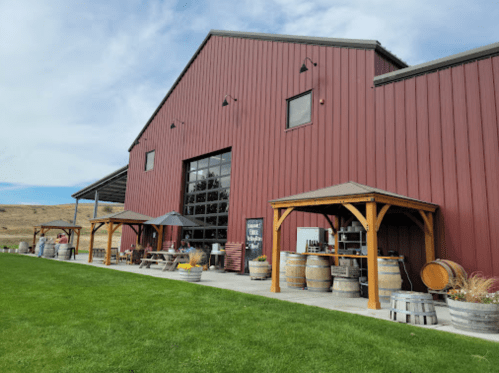 A red barn-style building with outdoor seating, wine barrels, and a grassy area under a blue sky.