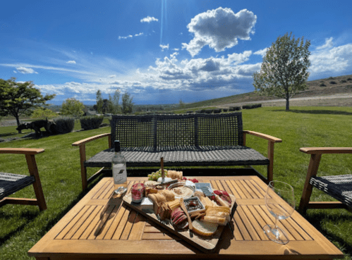 A picnic spread with wine and snacks on a wooden table, set in a sunny outdoor area with a scenic view.