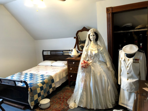 A vintage bedroom featuring a bed, dresser, and a mannequin in a wedding dress beside a military uniform.
