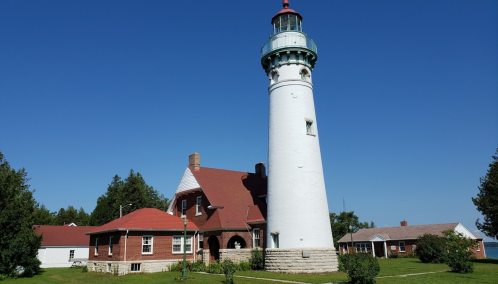 A tall white lighthouse with a glass lantern, next to a red-roofed building, set against a clear blue sky.