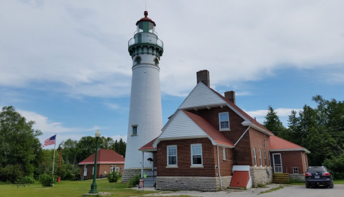 A tall white lighthouse beside a red-brick house, with trees and a flag in the background under a blue sky.