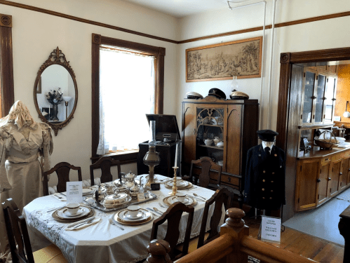 A vintage dining room with a table set for a meal, antique decor, and historical clothing displayed.