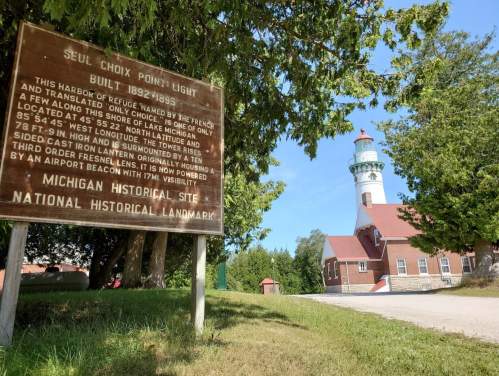 Sign for Seul Choix Point Light, with a lighthouse in the background surrounded by trees and a clear blue sky.