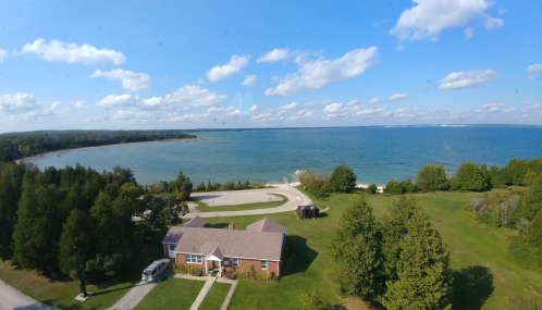 Aerial view of a house by a lake, surrounded by green trees and a clear blue sky with scattered clouds.