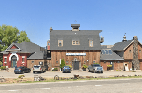 A rustic building with a wooden exterior and "ANTIQUE" sign, surrounded by cars and greenery under a clear blue sky.