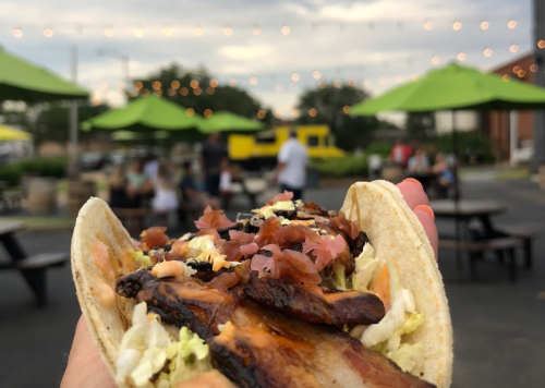 A hand holding a taco filled with meat, toppings, and slaw, with a lively outdoor setting in the background.