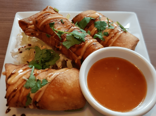 Three crispy spring rolls drizzled with sauce, served on a plate with a side of dipping sauce and garnished with cilantro.