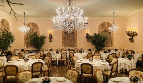 Elegant dining room with round tables, floral centerpieces, and a grand chandelier, featuring soft pink walls and draped windows.