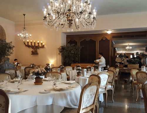 Elegant restaurant interior with chandeliers, round tables set for dining, and potted flowers on each table.