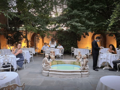 A charming outdoor restaurant with tables, a fountain, and guests dining under trees in soft evening light.