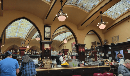 Interior of a bar with arched ceilings, stained glass, and patrons at the counter. Bottles line the shelves behind the bar.