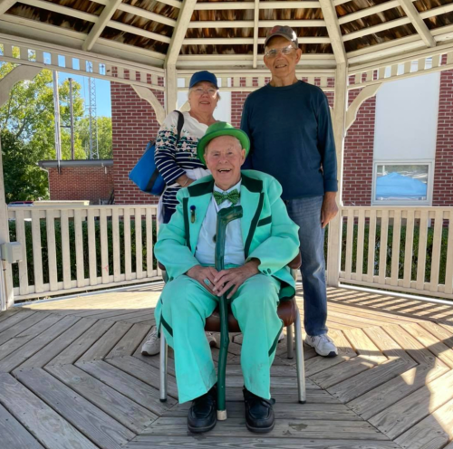 A man in a green suit sits in a gazebo, flanked by two smiling individuals, one wearing a blue hat and the other in glasses.