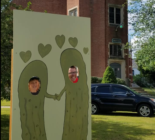 Two people with their faces in a pickle-themed cutout, holding hands with hearts above them, in a park setting.