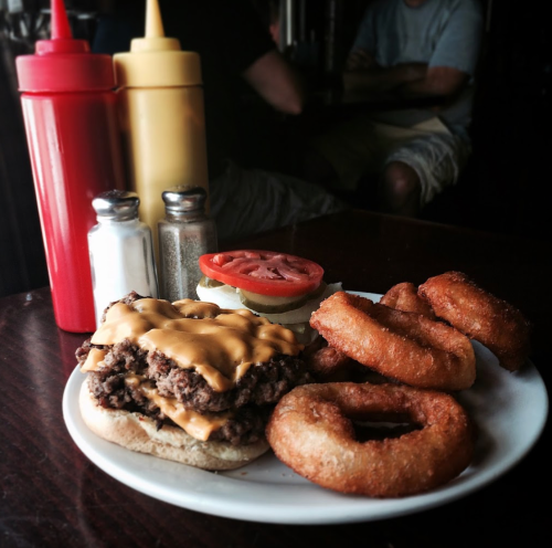 A plate with a cheeseburger topped with lettuce and tomato, accompanied by onion rings and condiments in the background.