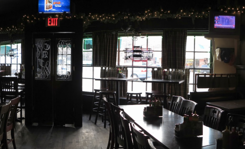 Interior of a dimly lit restaurant with wooden tables, chairs, and large windows, decorated with holiday lights.