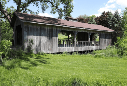 A rustic wooden covered bridge surrounded by lush green grass and trees under a clear blue sky.