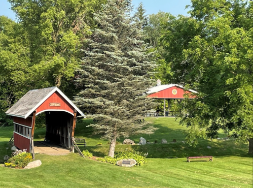 A red covered bridge beside a large evergreen tree, with a red building and green lawn in the background.