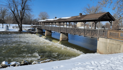 A snow-covered bridge spans a river, with flowing water and trees in the background under a clear blue sky.