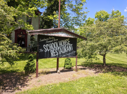 Sign for "The Schoolhouse Restaurant" surrounded by greenery, with a building visible in the background.