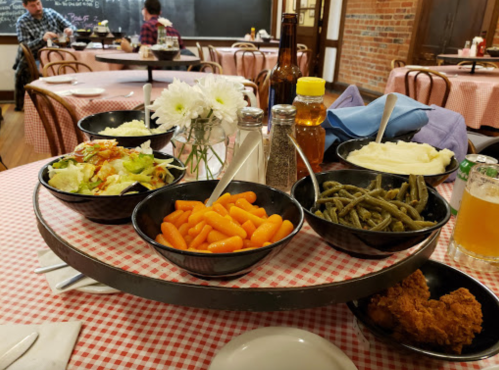 A round serving tray with bowls of carrots, green beans, mashed potatoes, salad, and fried chicken on a checkered tablecloth.