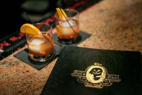 Two cocktails with orange slices on a bar counter, next to a black menu featuring a gold logo.