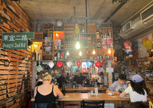 A cozy bar with wooden walls, shelves filled with bottles, and patrons enjoying drinks under warm lighting.