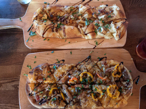 Two flatbreads on wooden boards, one topped with caramelized onions and the other with peppers and drizzled sauce.