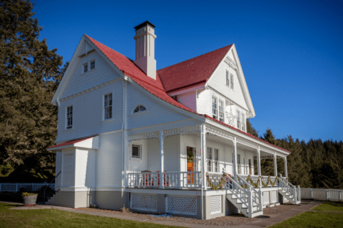 A charming white house with a red roof, featuring a porch and surrounded by trees under a clear blue sky.