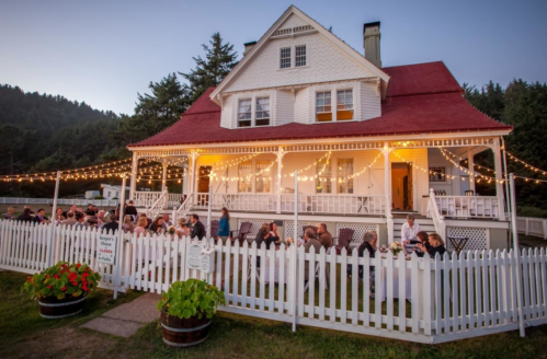 A charming white house with a red roof, decorated with string lights, hosts a dinner party on its porch.