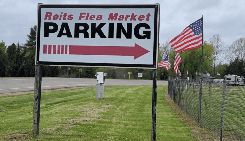 Sign for Reits Flea Market parking with an arrow pointing left, surrounded by American flags and green grass.