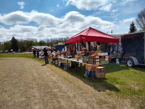A vibrant outdoor market scene with a red tent, tables of goods, and shoppers enjoying a sunny day.