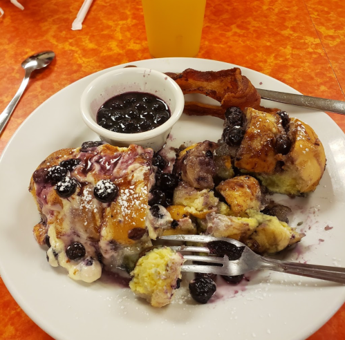 A plate with blueberry French toast, a side of syrup, and a piece of bacon, set on a bright orange table.