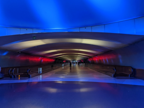 A long, illuminated corridor with blue and red lighting, featuring smooth floors and escalators on the sides.