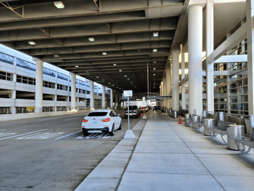 A spacious airport drop-off area with parked cars and seating along the sidewalk under a covered structure.