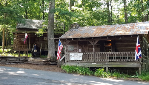 Two rustic log cabins surrounded by trees, featuring American and British flags, with a gravel road in front.