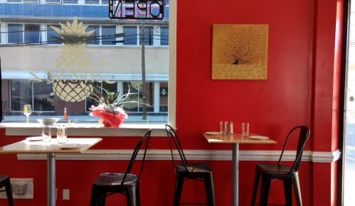 A cozy restaurant with red walls, two empty tables, and a window displaying a pineapple logo and a tree painting.