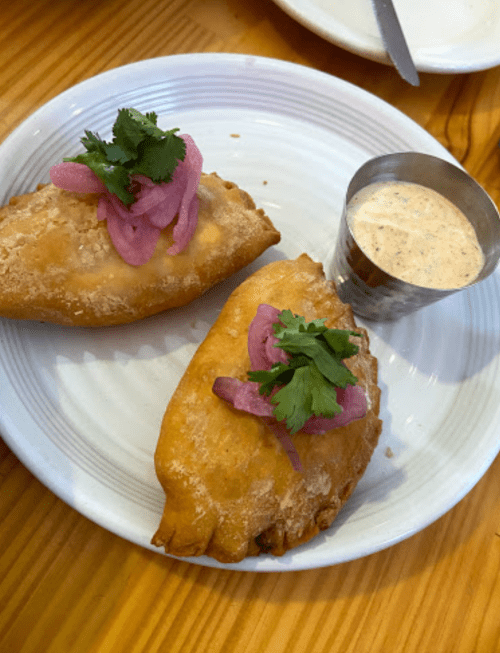 Two golden empanadas topped with pink onions and cilantro, served with a small cup of creamy dipping sauce on a white plate.