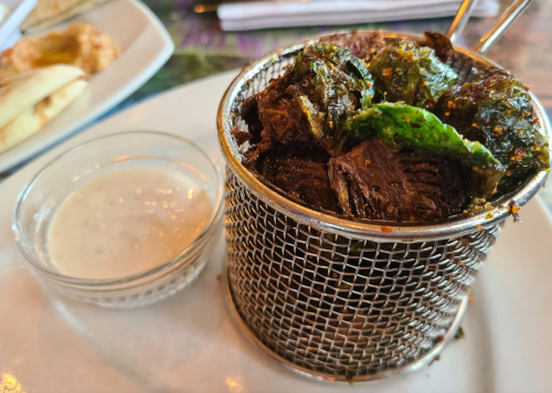 A small metal basket filled with crispy greens and a side of creamy dipping sauce on a white plate.