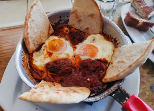 A skillet with two eggs cooked in a spicy sauce, surrounded by pieces of pita bread.