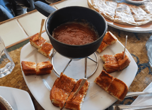 A pot of melted cheese surrounded by toasted bread pieces on a white plate, with a side of sauce.