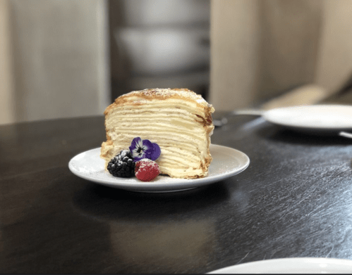 A slice of layered cake on a white plate, garnished with berries and a purple flower, on a dark wooden table.