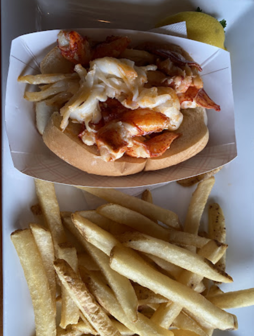 A lobster roll on a bun with fries on the side, garnished with a lemon wedge.