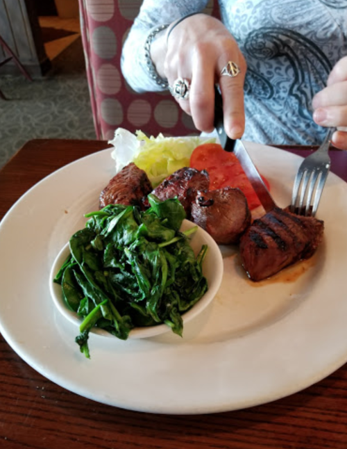 A plate with grilled steak, sautéed spinach, and a side of lettuce and tomato, with a person cutting the steak.