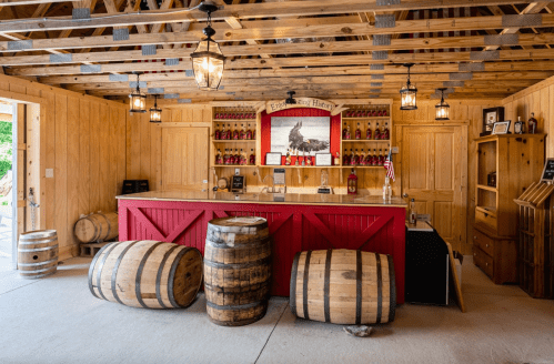 A rustic bar with wooden barrels, a red counter, and shelves filled with bottles in a warmly lit wooden interior.